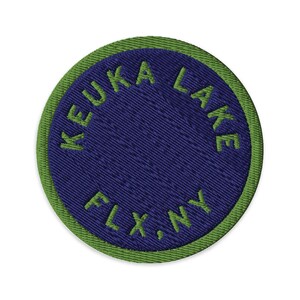 Könnte beinhalten: Ein kreisförmiger gestickter Aufnäher mit einer dunkelblauen Mitte und einem hellgrünen Rand. Der Text KEUKA LAKE ist in Grün entlang der oberen Kurve gestickt, und der Text FLX, NY ist in Grün entlang der unteren Kurve des Aufnähers gestickt.