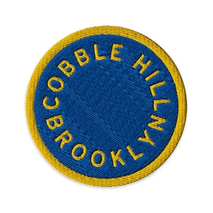 Może przedstawiać: Okrągła, haftowana naszywka z niebieskim środkiem i żółtą obwódką. Tekst "COBBLE HILL BROOKLYN" jest wyhaftowany na żółto wokół niebieskiego środka. Naszywka ma teksturowany wygląd.