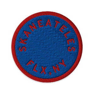 Könnte beinhalten: Ein kreisförmiger gestickter Aufnäher mit einer einfarbig blauen Mitte und einem roten Rand. Der Text SKANEATELES FLX, NY ist in rotem Garn kreisförmig entlang des inneren Randes des Aufnähers gestickt und erzeugt ein klares Design.