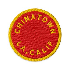 Może przedstawiać: Okrągła haftowana naszywka z żywym czerwonym środkiem i grubą żółtą obwódką. Napis CHINATOWN jest wygięty w łuk w górnej połowie, a napis L.A., CALIF. w dolnej połowie, oba wyhaftowane żółtą nicią na czerwonym tle naszywki.