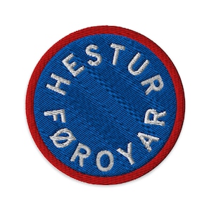 Puede incluir: Parche bordado redondo con centro azul y borde rojo. El centro azul presenta texto blanco en un patrón circular que dice "HESTUR FØRØYAR". El parche es redondo y tiene una apariencia texturizada.