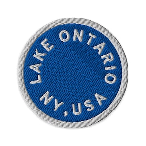 May include: A circular embroidered patch featuring a deep blue center with white stitched text reading LAKE ONTARIO NY, USA arranged in a circle. The patch has a thick, light gray stitched border around the entire outer edge of the design.