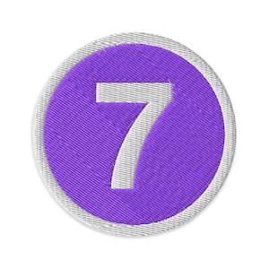 May include: Embroidered patch featuring the number 7 in white, centered on a purple circle. The circle is surrounded by a white stitched border. The patch has a textured appearance, suggesting it is made of fabric.