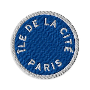 May include: A circular embroidered patch featuring a vibrant blue centre with the white text "ILE DE LA CITE PARIS" stitched in a circular pattern. The patch is finished with a thick, light grey stitched border around the entire circumference.