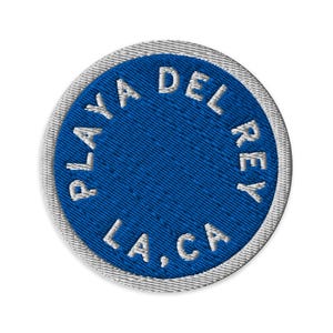 May include: A circular embroidered patch featuring a vibrant blue center with white stitched text that reads PLAYA DEL REY LA, CA arranged in a circular pattern. The patch is finished with a thick, light gray stitched border around the edge.