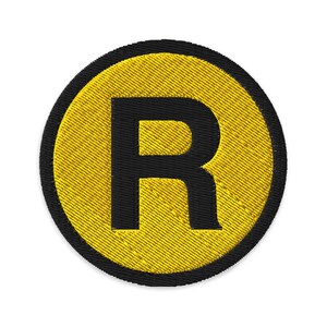 Pode incluir: Patch bordado com a letra "R" preta dentro de um círculo amarelo. O círculo amarelo tem uma aparência texturizada e é rodeado por uma borda preta. Ideal para personalizar jaquetas, bolsas ou outros artigos de tecido.