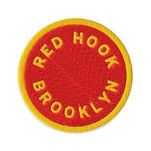 Pode incluir: Remendo bordado circular com centro vermelho e borda amarela. O texto "RED HOOK BROOKLYN" é bordado em amarelo, seguindo a forma do remendo. O remendo tem uma aparência texturizada.
