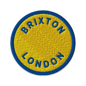 May include: A circular embroidered patch featuring a bright yellow center with a thick blue border. The text BRIXTON is stitched in blue along the top curve, and the text LONDON is stitched in blue along the bottom curve of the patch.