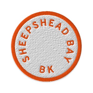 May include: A circular embroidered patch featuring a white centre with the orange text SHEEPSHEAD BAY BK arranged in a circular pattern, surrounded by a thick orange border. The embroidery has a visible diagonal stitch texture throughout.