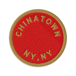 May include: A circular embroidered patch with a bright red center and a tan border. The text CHINATOWN is arched across the top half, and the text NY, NY is positioned in the bottom half, all rendered in tan, capitalized, sans-serif lettering.