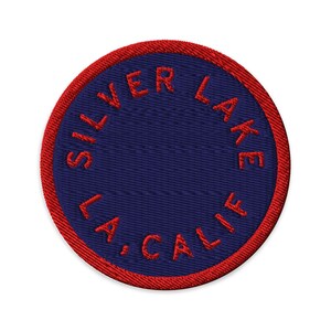 May include: A circular embroidered patch featuring a dark blue center and a vibrant red border. The text SILVER LAKE L.A., CALIF. is stitched in red thread in a circular pattern around the inner edge of the patch, creating a bold contrast.