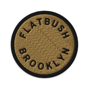 Può includere: Una toppa ricamata circolare con sfondo color marrone chiaro e un bordo nero spesso. Il testo "FLATBUSH BROOKLYN" è ricamato in lettere maiuscole nere, seguendo la curva circolare della toppa lungo il bordo interno della cornice.