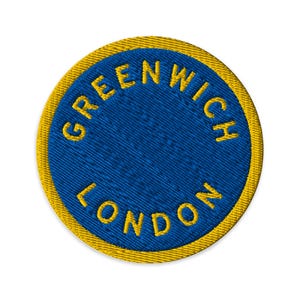 May include: A circular embroidered patch featuring a deep blue center and a thick yellow border. The text GREENWICH LONDON is stitched in yellow capital letters along the inner edge of the border, following the circular shape of the patch.