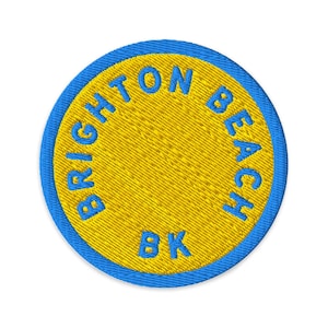 Może przedstawiać: Okrągła haftowana naszywka z jaskrawożółtym środkiem i niebieską obwódką. Napis BRIGHTON BEACH BK jest wyhaftowany niebieską nicią w okrągły wzór wokół środka naszywki, przy czym BK znajduje się na dole.