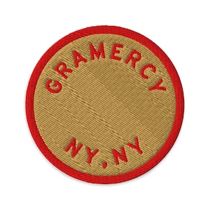 Pode incluir: Um emblema bordado circular com um centro bege de textura de sarja diagonal e uma borda vermelha vibrante. O texto GRAMERCY está arqueado no topo e o texto NY. NY. está posicionado na parte inferior, tudo bordado com fio vermelho.