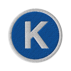 Puede incluir: Parche bordado con la letra "K" blanca sobre fondo azul. La letra está centrada en un diseño circular, rodeada por un borde gris claro. El parche tiene una apariencia texturizada.