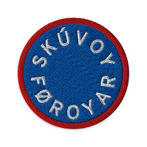 May include: Embroidered circular patch with a blue center and a red border. The blue center features white text that reads "SKÚVOYAR FØROYAR" in a circular pattern. The patch is likely for apparel or accessories.