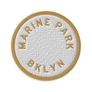 Puede incluir: Un parche bordado circular con un centro blanco y el texto dorado MARINE PARK BKLYN cosido en un patrón circular. El parche tiene un borde dorado grueso y un acabado de tela texturizada, diseñado para su aplicación en ropa o accesorios.