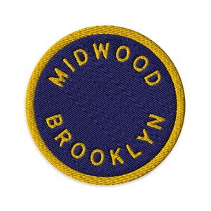 Puede incluir: Un parche bordado circular con un centro azul oscuro y un borde amarillo grueso. El texto MIDWOOD BROOKLYN está cosido con hilo amarillo, curvándose a lo largo de los bordes superior e inferior del campo circular azul con un acabado texturizado.
