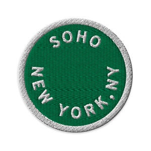 May include: A circular embroidered patch featuring a deep green background and a white border. The text SOHO NEW YORK is stitched in white capital letters, arranged in a circular pattern along the inner edge of the patch's green field.