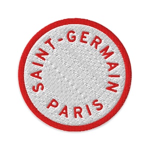 May include: A circular embroidered patch featuring a white center and a thick red border. The text SAINT-GERMAIN PARIS is stitched in red capital letters along the inner edge of the border, following the circular shape of the patch.