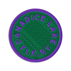 Könnte beinhalten: Ein kreisförmiger gestickter Aufnäher mit einer leuchtend grünen Mitte und einem violetten Rand. Der Text CANADICE LAKE FLX, NY ist in violettem Garn entlang des inneren Randes gestickt und umschließt das einfarbige grüne kreisförmige Feld.