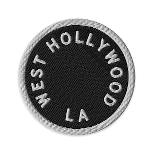 Może przedstawiać: Okrągła haftowana naszywka z czarnym tłem i białą obwódką. Napis WEST HOLLYWOOD LA jest wyhaftowany białymi wielkimi literami w okrągły wzór wokół środka naszywki, co tworzy przejrzysty i wyrazisty projekt graficzny.