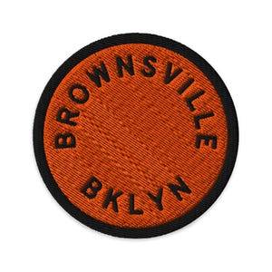 Puede incluir: Un parche bordado circular con un fondo naranja vibrante y un borde negro grueso. El texto BROWNSVILLE está arqueado en la mitad superior y el texto BKLYN está centrado en la parte inferior, ambos bordados con hilo negro grueso.