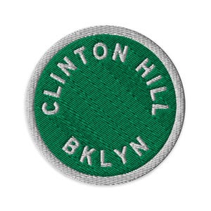 Op de afbeelding: Een ronde geborduurde patch met een felgroen midden en de witte tekst CLINTON HILL en BKLYN in een cirkelvormig patroon gestikt. De patch is afgewerkt met een witte geborduurde rand rondom de gehele buitenste rand.