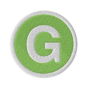 Puede incluir: Parche bordado circular con la letra "G" verde dentro de un contorno blanco y un borde verde. El parche tiene un aspecto texturizado, adecuado para aplicar en ropa o accesorios.