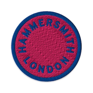 May include: A circular embroidered patch featuring a vibrant magenta center and a deep blue border. The text HAMMERSMITH LONDON is stitched in a bold blue font, curving along the inner edge of the patch, creating a clean and structured design.