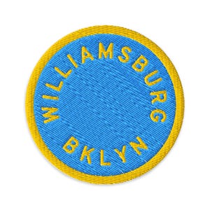 May include: Embroidered patch with a blue center and a yellow border. The text "WILLIAMSBURG BKLYN" is embroidered in yellow around the blue circle. The patch is circular and has a textured appearance.