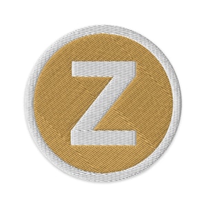 Può includere: Toppa ricamata con la lettera "Z" bianca, centrata su un cerchio color oro. Il cerchio è circondato da un bordo bianco. La toppa ha un aspetto strutturato, suggerendo che sia fatta di tessuto.