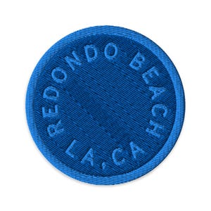 Può includere: Una toppa circolare blu ricamata con un bordo cucito. Il testo REDONDO BEACH LA, CA è impresso in un motivo circolare attorno al centro della toppa, con le lettere e lo sfondo realizzati nella stessa tonalità di colore blu.