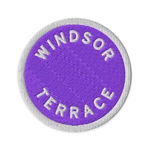 May include: A circular embroidered patch featuring a vibrant purple centre with the white text WINDSOR TERRACE stitched in a curved pattern along the top and bottom edges, finished with a white stitched border around the entire circumference.