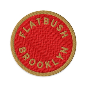 May include: A circular embroidered patch featuring a vibrant red center with the text FLATBUSH BROOKLYN stitched in gold thread along the inner perimeter. The patch is finished with a thick, textured gold border that frames the red circular design.