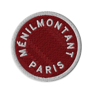 May include: A circular embroidered patch featuring a deep red center and a white border. The text MENILMONTANT PARIS is stitched in white capital letters in a circular pattern along the inner edge of the red field, creating a clean design.