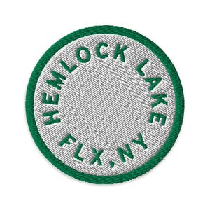 Könnte beinhalten: Ein kreisförmiger gestickter Aufnäher mit weißer Mitte und einem dicken grünen Rand. Der Text "HEMLOCK LAKE FLX, NY" ist in grünem Garn kreisförmig entlang des inneren Randes der Umrandung gestickt und umschließt die weiße Mitte.