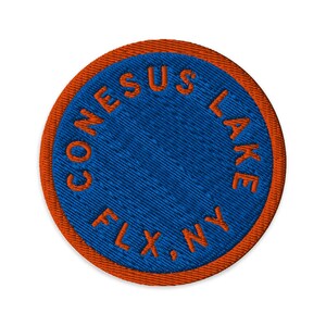 Könnte beinhalten: Ein kreisförmiger bestickter Aufnäher mit einer leuchtend blauen Mitte und einem kräftigen orangefarbenen Rand. Der Text CONESUS LAKE, NY ist in orangefarbenem Garn kreisförmig entlang des inneren Randes gestickt und sorgt für ein klares Design.