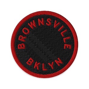 May include: A circular embroidered patch featuring a black centre and a red border. The text BROWNSVILLE BKLYN is stitched in red capital letters along the inner edge of the circular border, creating a bold contrast against the black background.