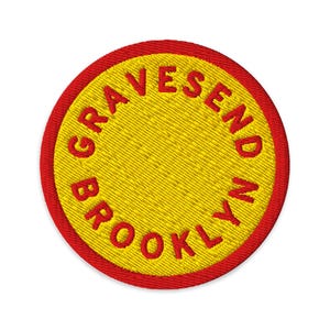 Puede incluir: Un parche bordado circular con un centro amarillo brillante rodeado por un borde rojo intenso. El texto GRAVESEND BROOKLYN está bordado en letras mayúsculas rojas siguiendo la curva circular del parche, creando un emblema distintivo.