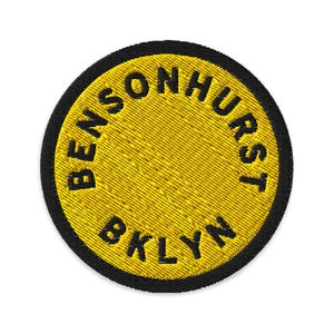 Puede incluir: Un parche bordado circular con un fondo amarillo brillante y un borde negro grueso. El texto BENSONHURST BKLYN está bordado en letras mayúsculas negras, siguiendo la curva circular del parche a lo largo del borde exterior.