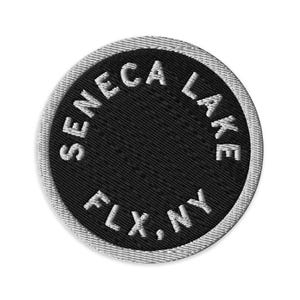 Könnte beinhalten: Ein runder gestickter Aufnäher mit schwarzer Mitte und weißem Sticktext, der kreisförmig SENECA LAKE FLX, NY lautet. Der Aufnäher hat einen dicken, hellgrau gestickten Rand, der das schwarze Innendesign deutlich umrahmt.