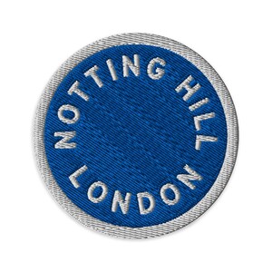 May include: A circular embroidered patch featuring a vibrant blue centre with the white text NOTTING HILL LONDON stitched in a circular pattern along the edge. The patch is finished with a thick, light grey stitched border around the circumference.