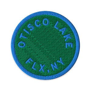 Könnte beinhalten: Ein kreisförmiger gestickter Aufnäher mit einer leuchtend grünen Mitte, die von einem hellblauen Rand umgeben ist. Der Text OTISCO LAKE ist in Blau entlang der oberen Kurve gestickt, mit FLX, NY in Blau entlang der unteren Kurve des Aufnähers.