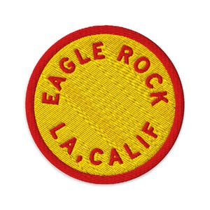 May include: A circular embroidered patch featuring a bright yellow center with a thick red border. The text EAGLE ROCK is arched across the top and LA, CALIF is arched across the bottom in bold red embroidered lettering against the yellow background.