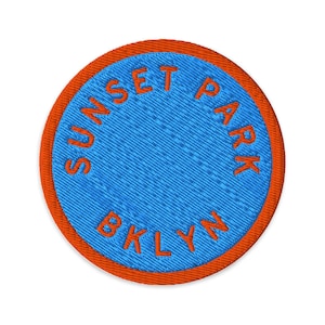 Op de afbeelding: Een ronde geborduurde patch met een felblauw midden en een dikke oranje rand. De tekst SUNSET PARK BKLYN is in oranje draad in een cirkelvormig patroon langs de binnenrand van de patch geborduurd, wat zorgt voor een opvallend contrast.