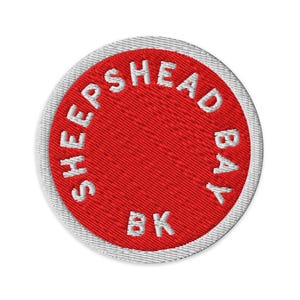 Op de afbeelding: Een cirkelvormige geborduurde patch met een felrood midden en een witte rand. De witte tekst SHEEPSHEAD BAY BK is in een cirkelvormig patroon langs de binnenrand van het rode veld geborduurd, met een textuur van draad over het geheel.