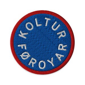 Op de afbeelding: Geborduurde ronde patch met een blauw midden en een rode rand. De witte tekst "KULTUR FØROYAR" is rond het midden geborduurd. Deze patch is ontworpen om op kleding of accessoires te worden genaaid.