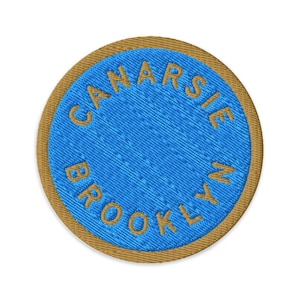 Puede incluir: Un parche bordado circular que presenta un centro azul brillante con el texto CANARSIE BROOKLYN cosido en hilo dorado. El parche tiene un borde dorado grueso que enmarca el área circular azul, creando un diseño limpio y definido.